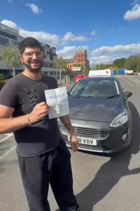 A bearded man in glasses holding a document in front of a grey car on a sunny street