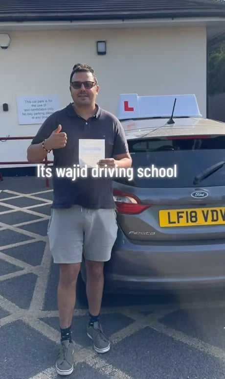 Man giving a thumbs up in front of a driving school car with an L-plate in a parking lot