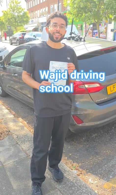 Young man holding a driving certificate standing next to a car with Wajid Driving School overlay
