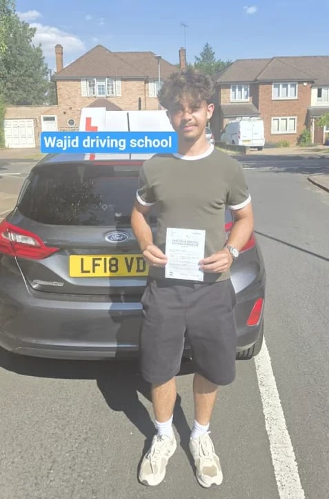 Student holding a driving certificate in front of a car with the Wajid Driving School sign